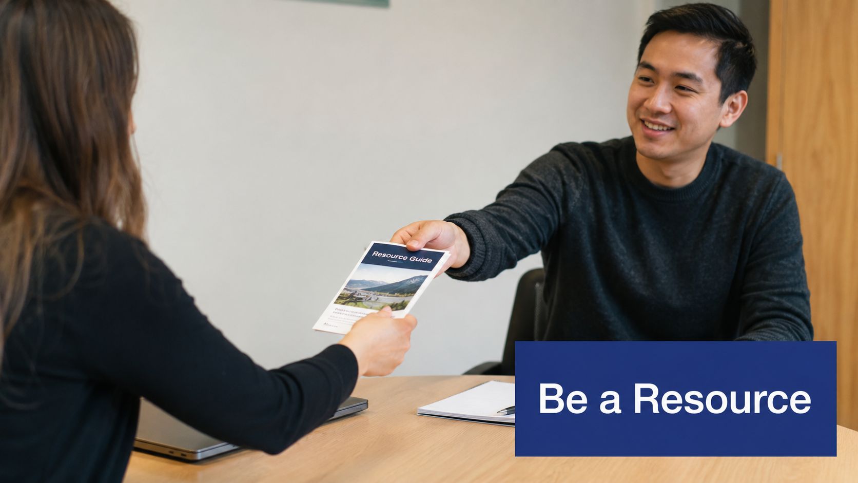 A professional man handing a printed resource guide to a woman across a desk in an office.